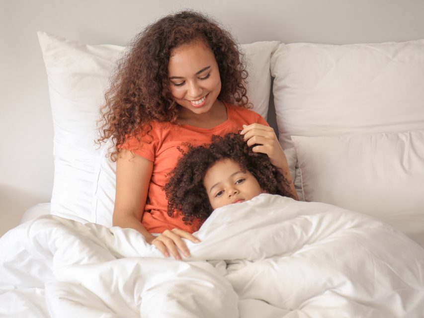 A child lying in bed under a white duvet, a parent sitting beside her