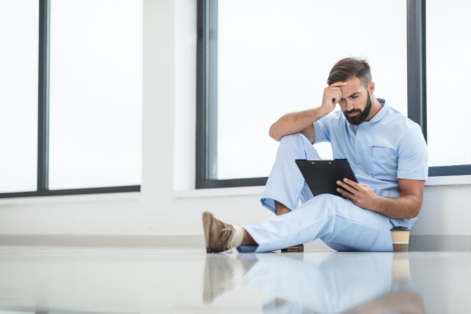 A clinician in scrubs sitting on the floor against a window, head in hand, clipboard on his knee