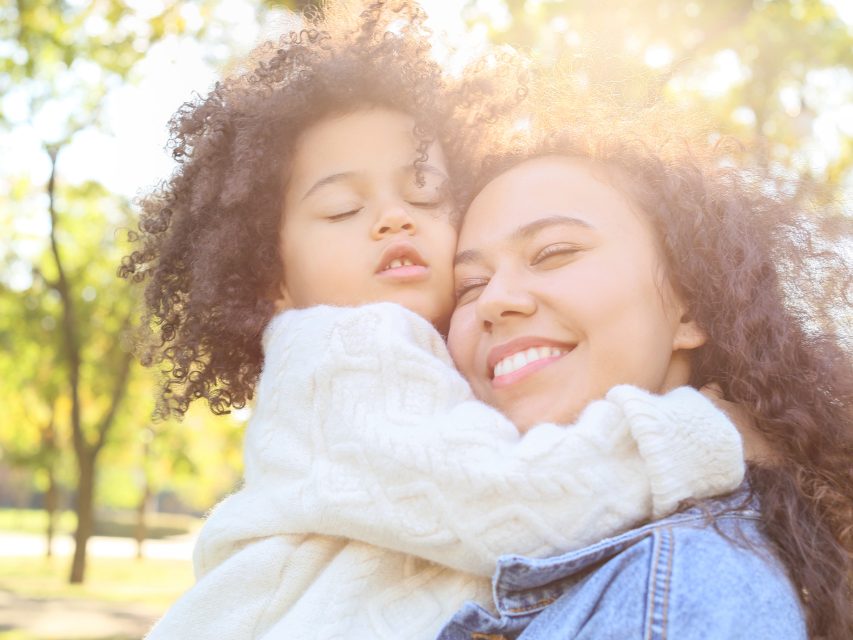 A mother holding her daughter close, both smiling, outdoors in soft light